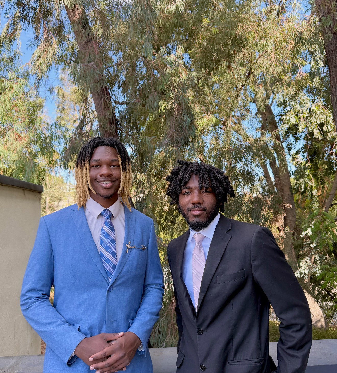 Two young businessmen with stylish dreadlock and twist hairstyles wearing blue and black suits, posing outdoors for a professional networking or entrepreneurship feature.