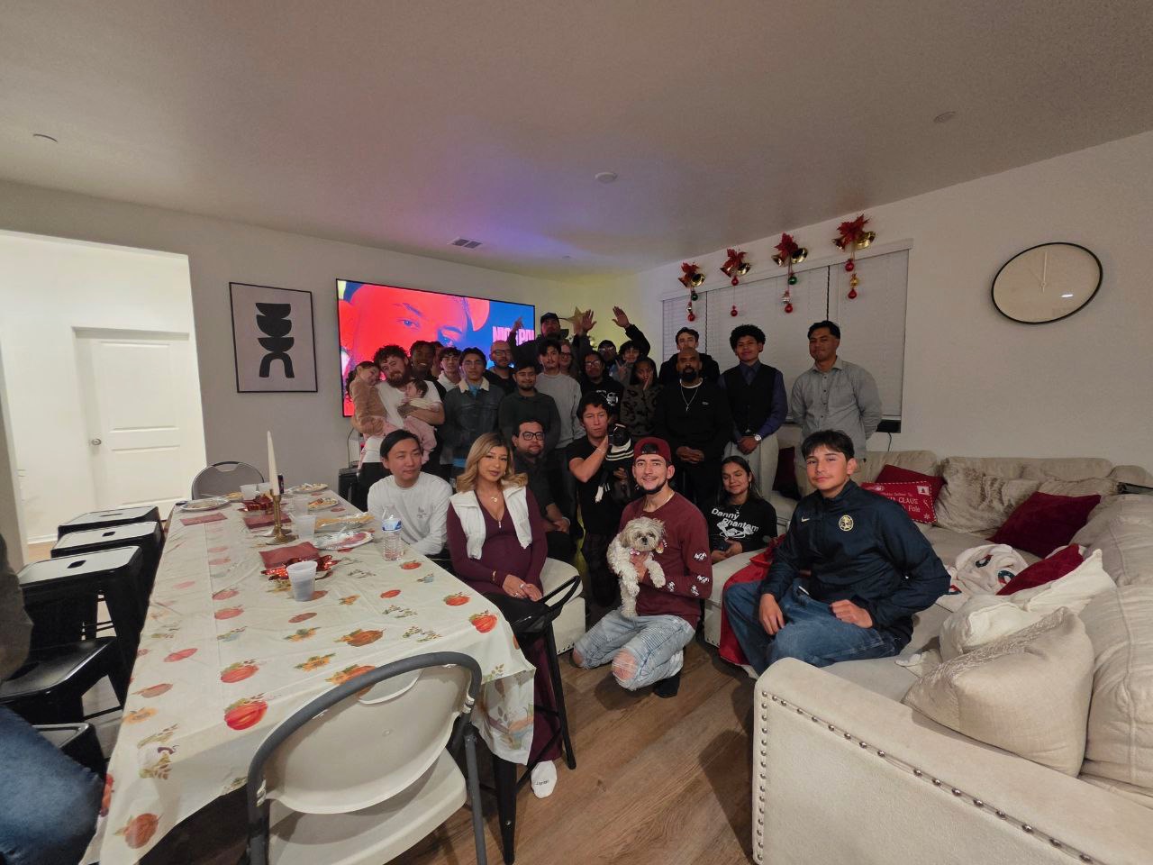 A large, diverse group of employees and family members gathered in a decorated living room for a corporate holiday party and team-building celebration.