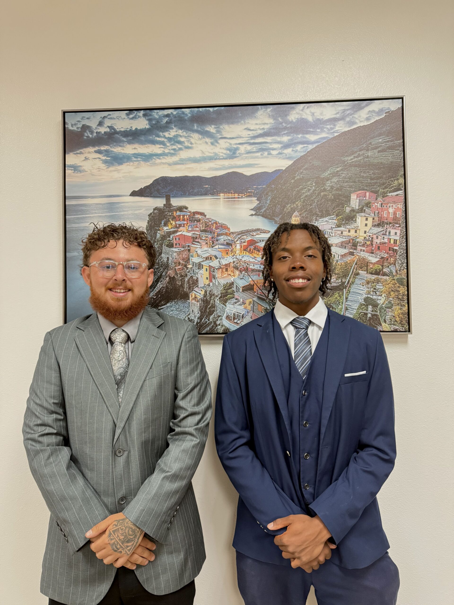 Two young professional businessmen smiling in grey and navy blue suits standing in front of a coastal landscape painting in a modern office setting.